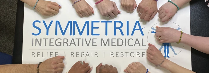 Group displaying joined hands around a Symmetria Integrative Medical sign promoting relief, repair, and restore.